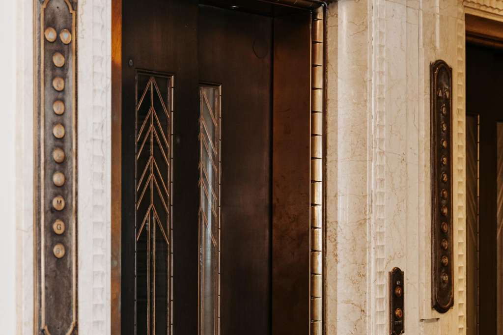 Ornate elevator doors with art deco detailing and brass call buttons in a marble-clad corridor.
