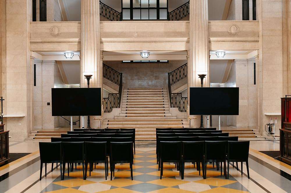 Main Hall set up with rows of black chairs facing two screens and a grand staircase, classical architecture with columns.