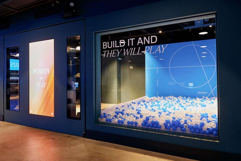 Exhibition display with blue and white ball pit installation, illuminated signage reading 'BUILD IT AND THEY WILL PLAY' on dark blue walls.