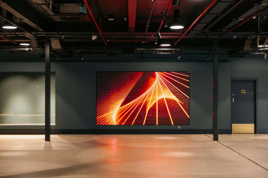 Industrial exhibition space with polished concrete floor, exposed red pipework, and large digital display screen.