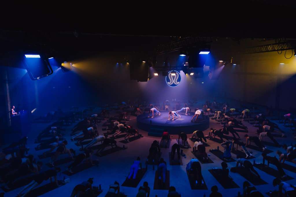 Large indoor space with blue and yellow stage lighting, performers on central platform, and many people exercising on mats.