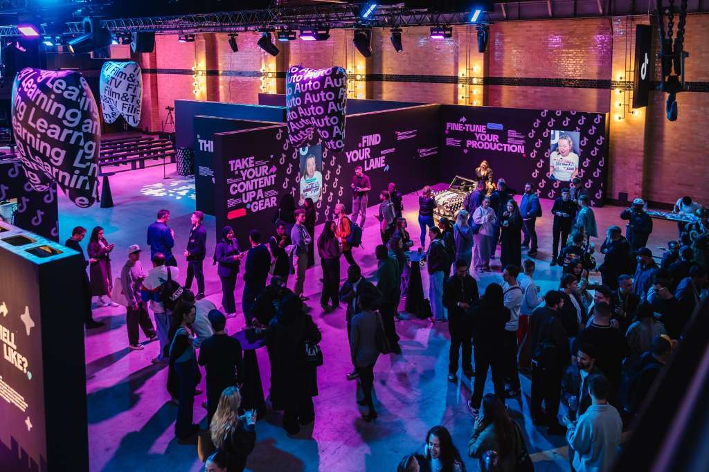 Large exhibition space with vibrant pink and blue lighting, multiple display stands, and attendees mingling throughout.