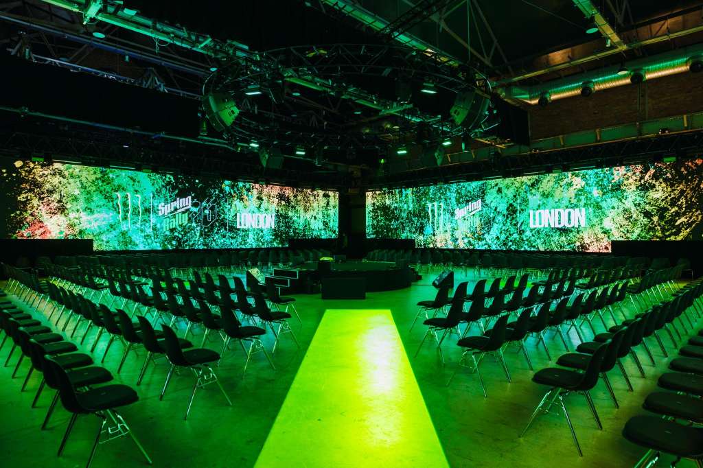 Large auditorium with rows of black chairs facing dual projection screens displaying green foliage and text, illuminated by green stage lighting.