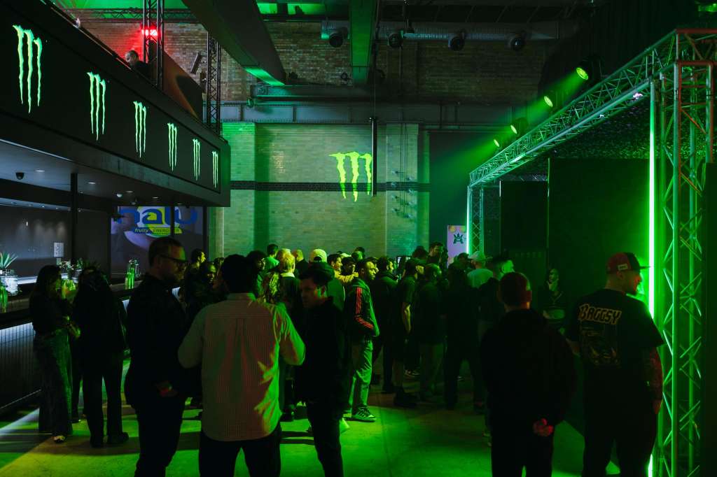 Industrial venue with green neon lighting, branded signage, and crowd gathered for an event.