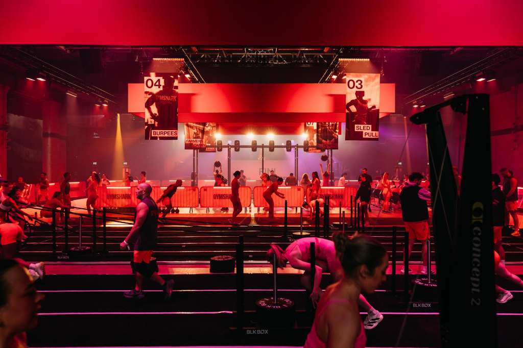 Large exhibition space with stage, dramatic red lighting, and participants engaged in team activities and fitness demonstrations.