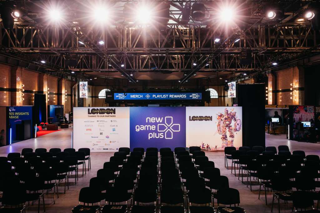 Large exhibition hall with rows of black chairs facing a stage displaying branded banners and sponsor logos under bright overhead lighting.