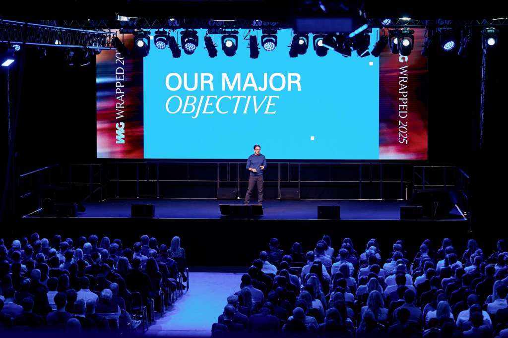 Large auditorium with audience seated facing a stage. Presenter stands under a bright blue projection screen with professional lighting rig overhead.