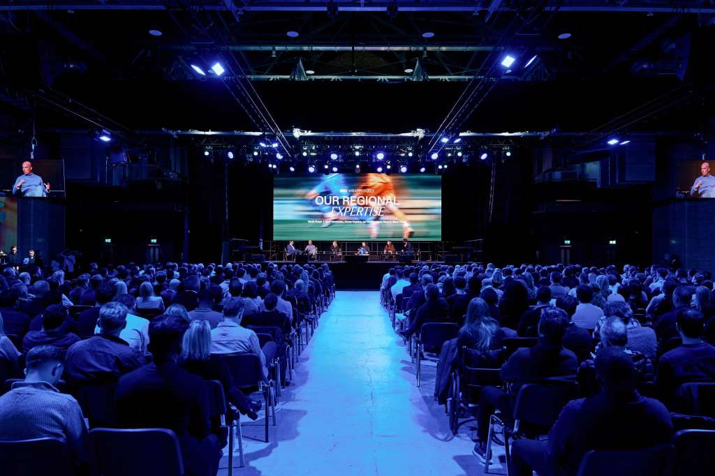 Large auditorium with rows of seated attendees facing a stage with projection screen and panel speakers under blue lighting.