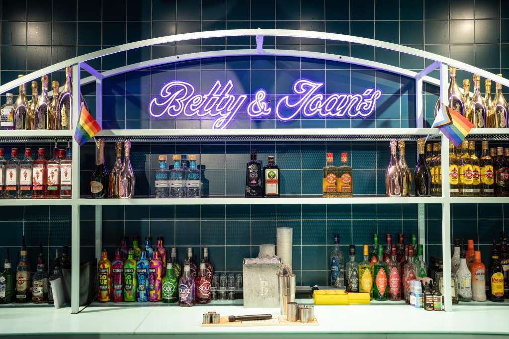 Betty & Joan's Main Space bar counter with neon signage, tiered bottle display, and pride flags.
