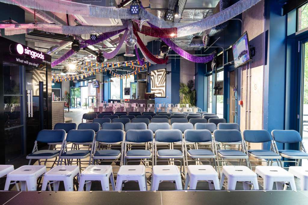 Main space set up with rows of blue chairs and white stools facing a stage, decorated with purple and red streamers and professional lighting.