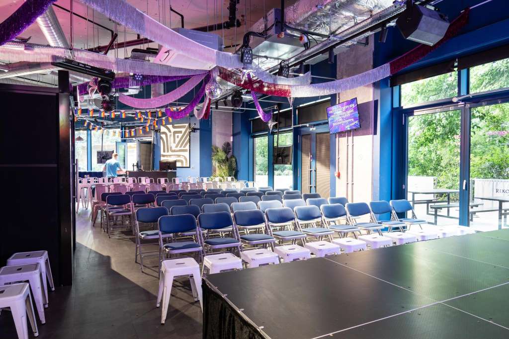 Modern event space with rows of blue chairs, purple and blue ambient lighting, stage area with bar, and large windows overlooking greenery.
