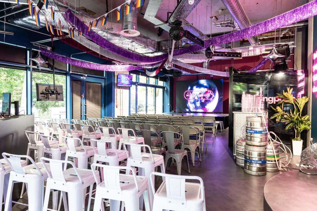 Modern event space with rows of white and pink chairs, purple neon lighting, decorative ribbons, and a large screen display.