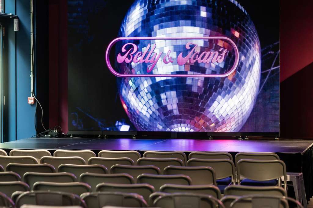 Main space with stage, large disco ball, purple and blue lighting, sequined draping, and rows of white chairs.