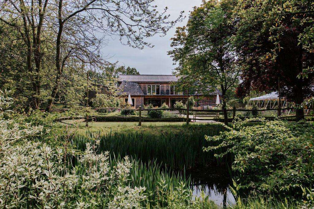 Modern brick building with large windows set in landscaped grounds, surrounded by mature trees and a pond with reeds.