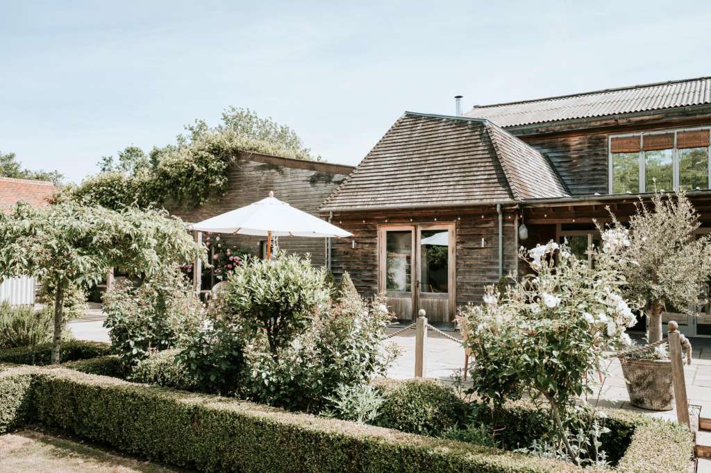 Exterior terrace with white flowering shrubs, parasol, and wooden barn building with manicured hedging.