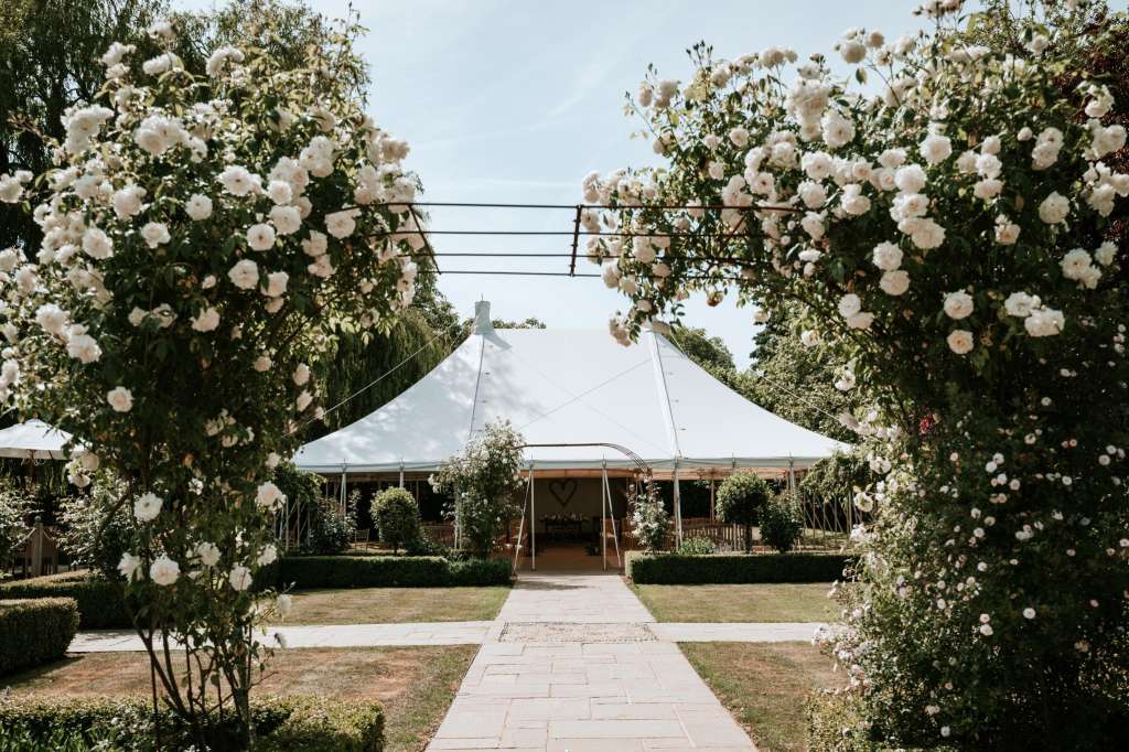 Exterior garden pathway lined with white flowering roses leading to a marquee tent with clear roof.