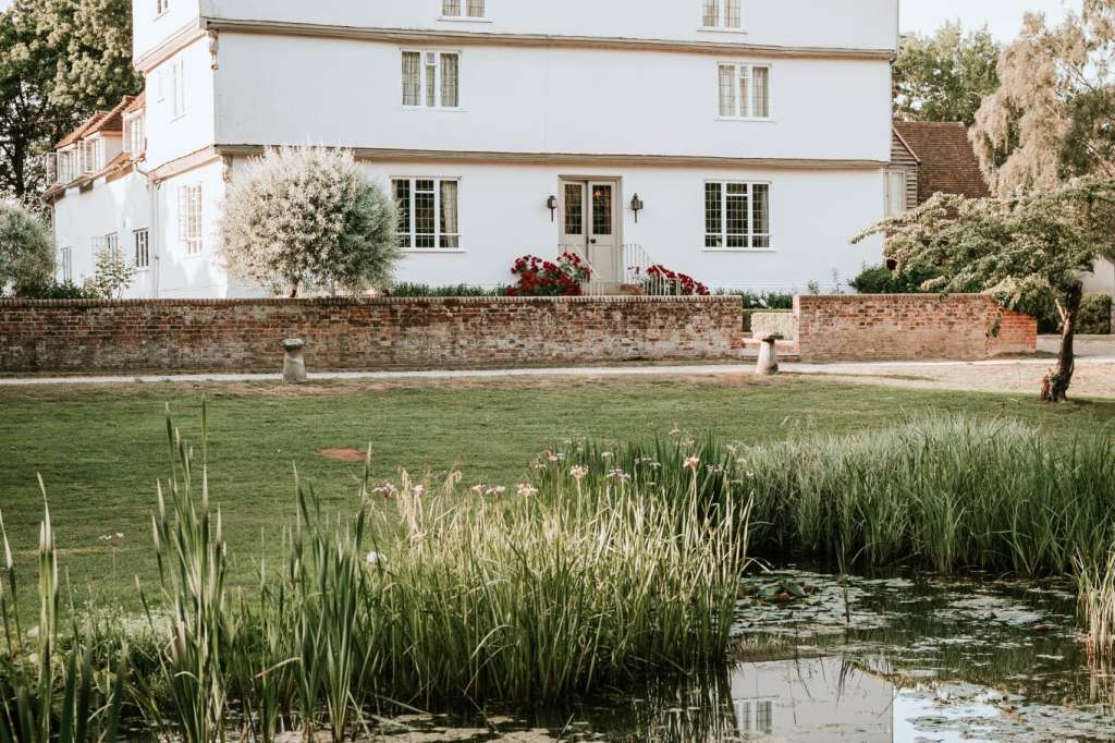 White historic manor house with brick chimney viewed across a pond with reeds and manicured lawn.