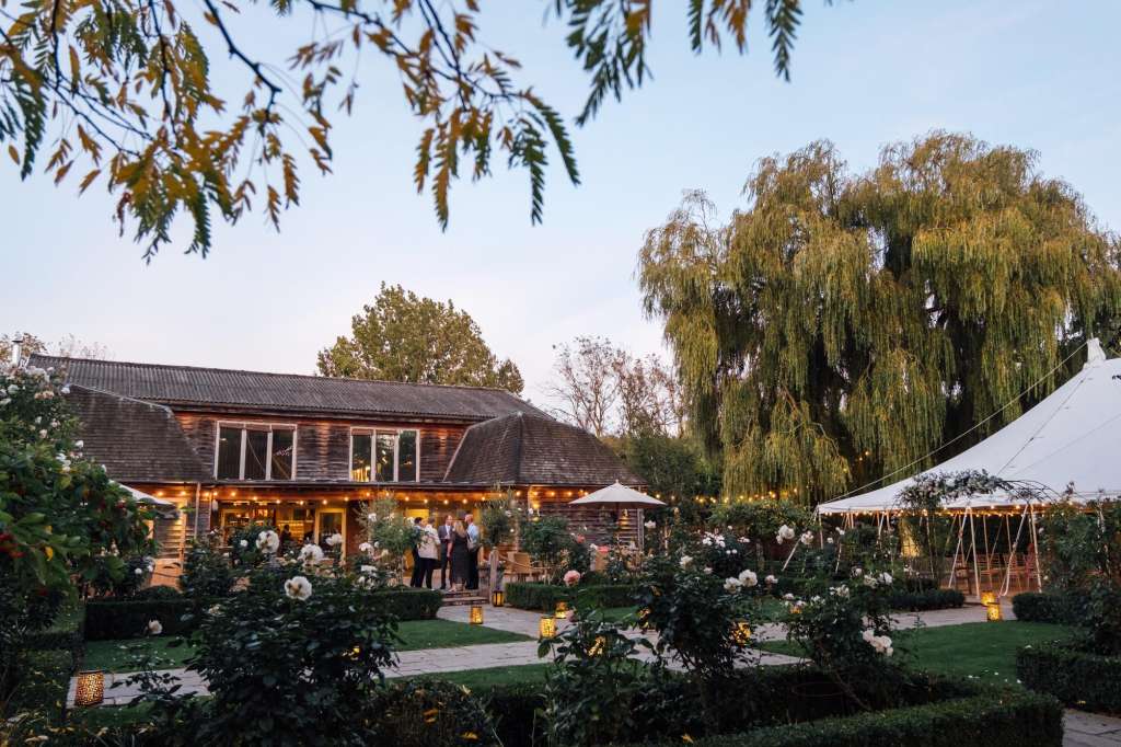 Outdoor evening reception with illuminated barn, white marquees, flowering plants, and guests mingling on paved terrace.