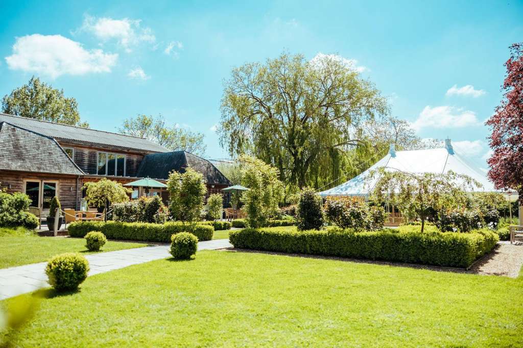 Exterior grounds with manicured lawns, hedges, weeping willow tree, and white marquee tent beside converted barn building.