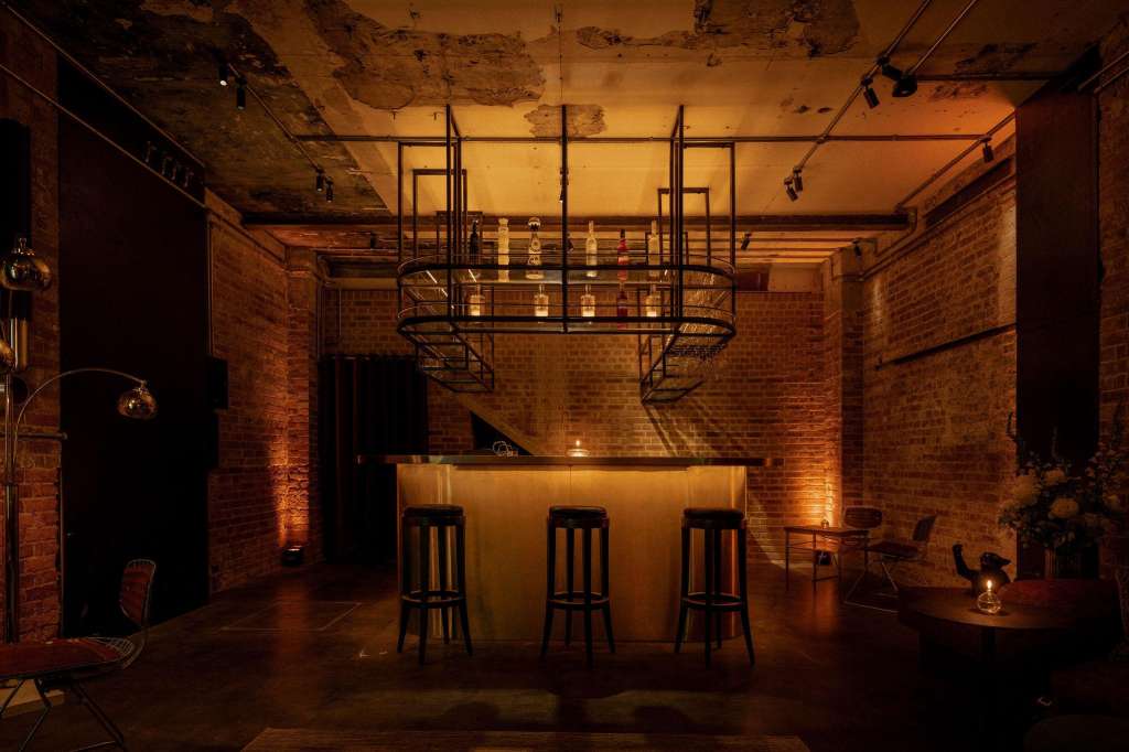 Industrial studio with exposed brick, bar counter, bar stools, and suspended metal shelving under warm lighting.