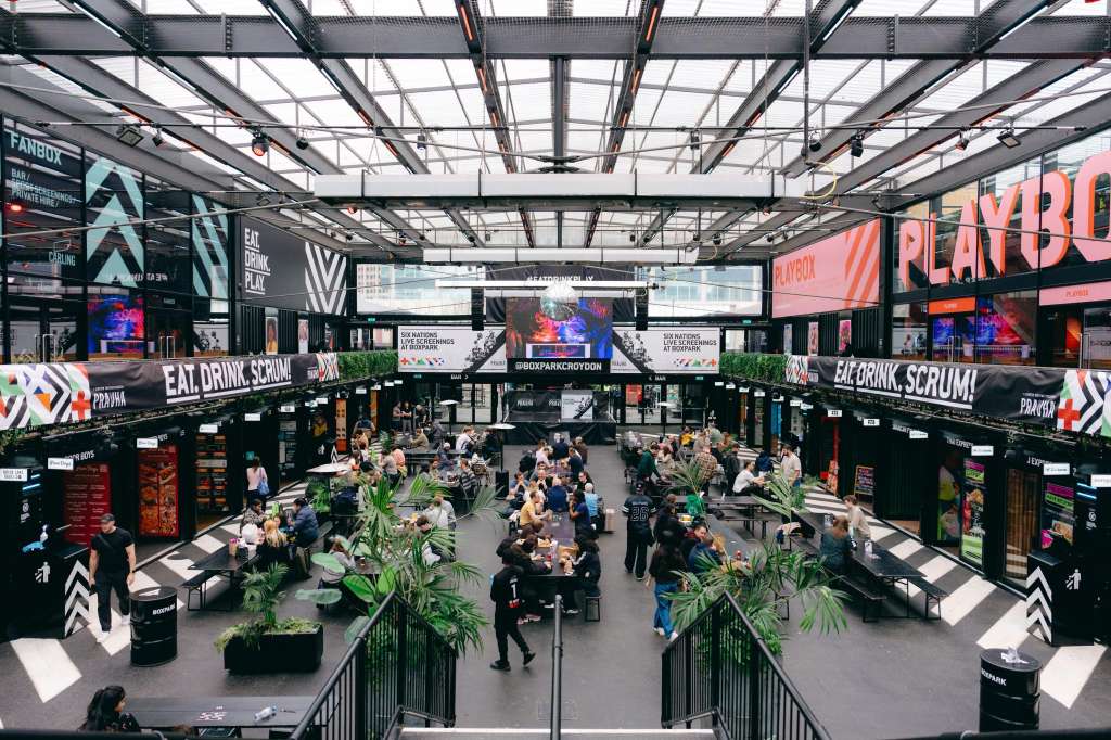 Modern food hall with industrial ceiling, black vendor stalls, digital screens, and crowds of people dining and socializing.