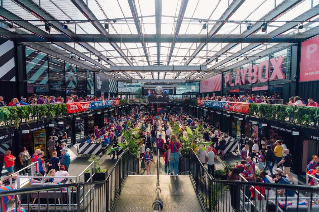 Large indoor marketplace with glass roof, multiple vendor stalls, crowds of visitors, and branded signage.