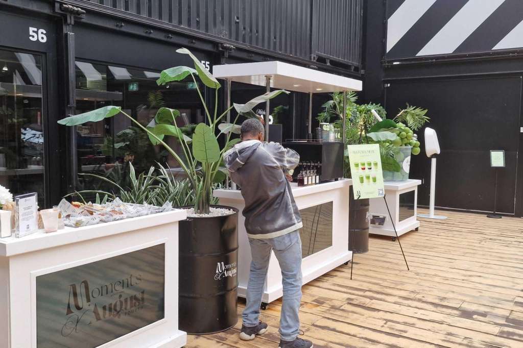 Outdoor rooftop space with bar counter, potted plants, and wooden decking. Modern industrial architecture with black cladding.