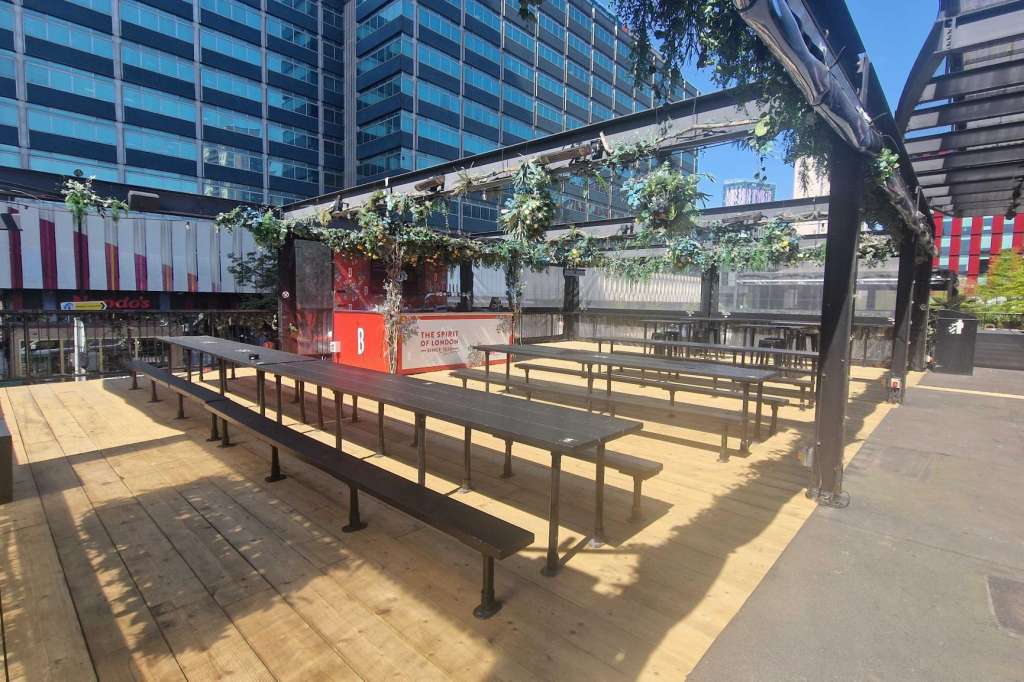 Outdoor rooftop terrace with wooden benches, pergola shade structure, red and white branding, and green climbing plants.