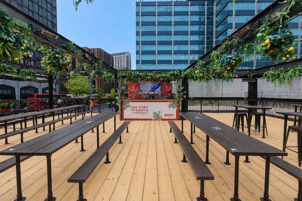 Outdoor rooftop terrace with wooden decking, long benches, lush green pergola, and stage setup with branded signage.