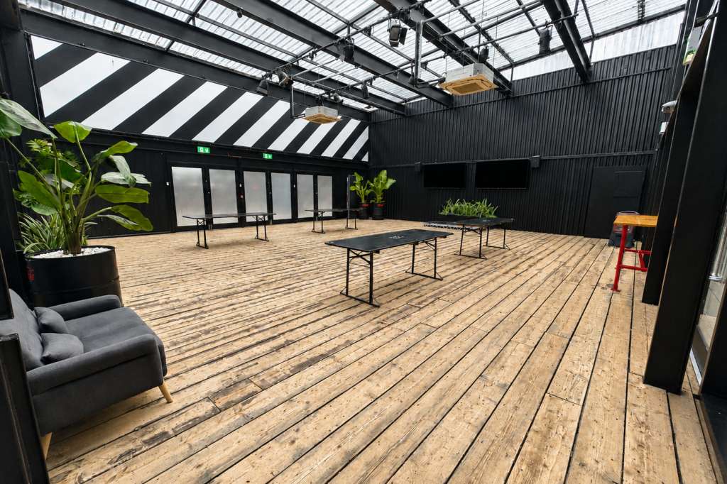 Industrial warehouse space with wooden flooring, black steel frame, skylights, ping-pong table, and potted plants.