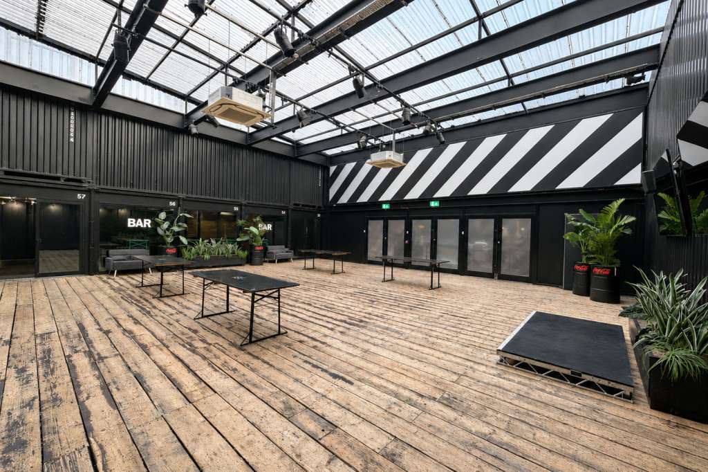 Industrial rooftop space with polished wood flooring, glass roof panels, black tables, and potted plants.