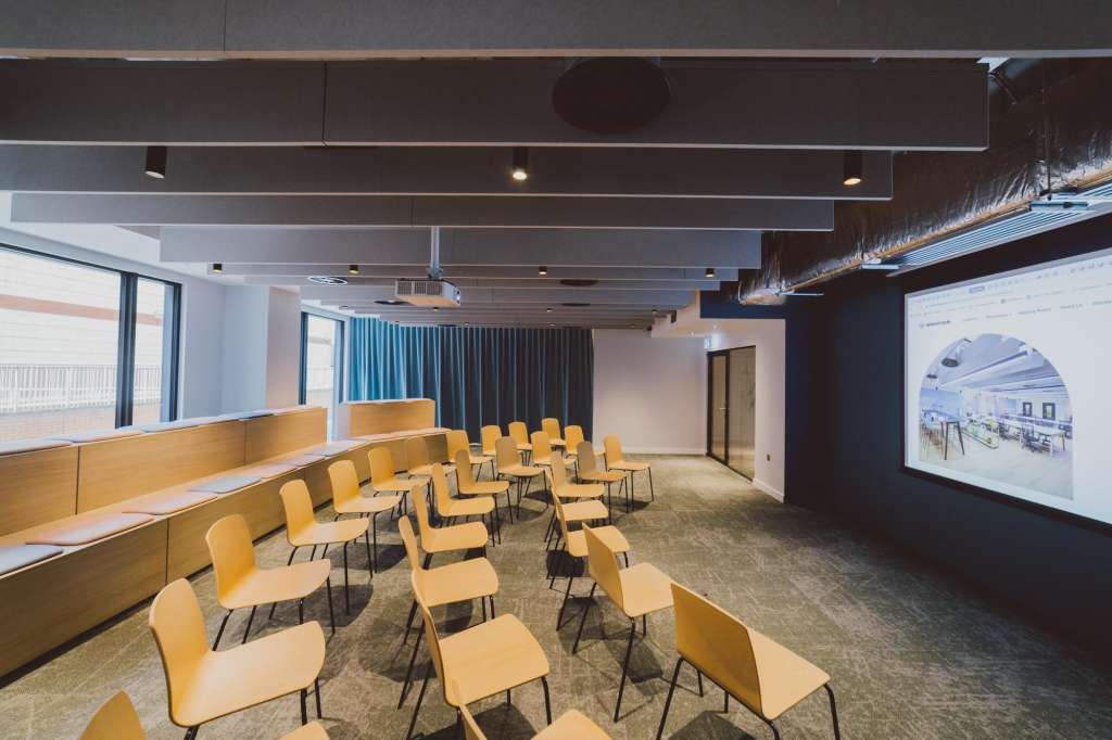 Modern conference room with yellow chairs arranged in rows, projection screen, and floor-to-ceiling windows.