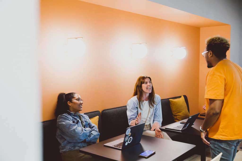 Modern office space with three people collaborating around a table with laptops, warm orange accent wall and recessed lighting.
