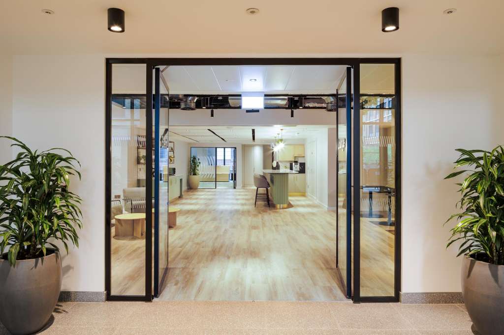 Modern open-plan office space with light wood flooring, black-framed glass partitions, potted plants, and recessed lighting.