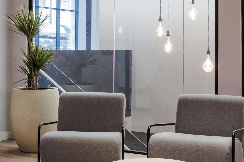 Modern lounge area with gray upholstered chairs, round wooden tables, pendant lighting, and potted plants near floor-to-ceiling windows.