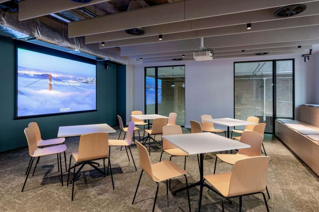Modern meeting room with multiple tables, wooden chairs, large projection screen displaying Zoom interface, and floor-to-ceiling windows.