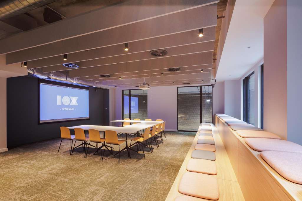 Modern meeting room with projection screen, rectangular table, orange chairs, and bench seating along windows.