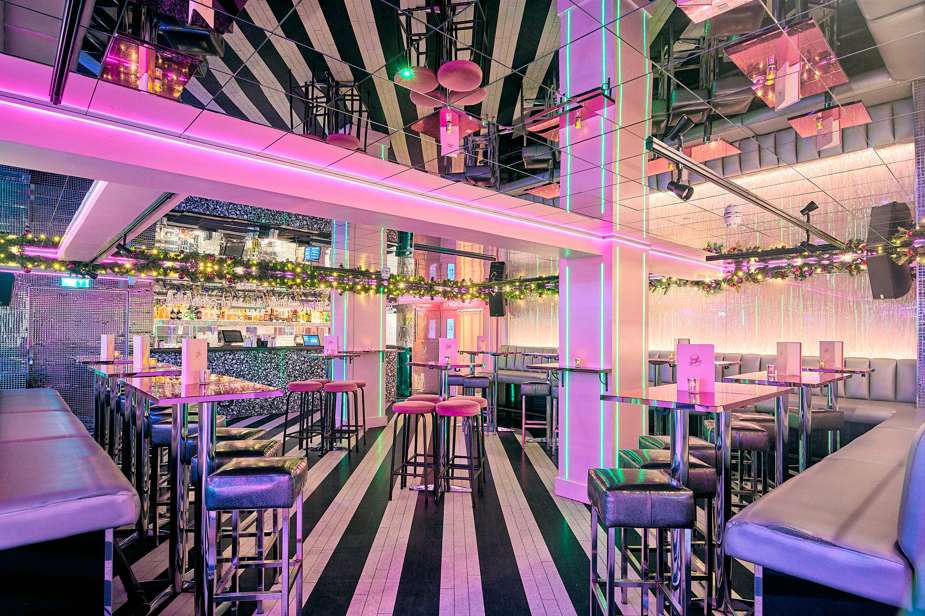 Modern venue interior with striped ceiling, neon pink and green lighting, bar counter, high-top seating, and geometric decor.