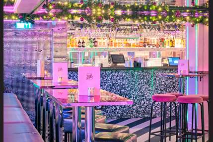 Modern bar interior with neon pink lighting, high-top seating, tiered shelving with bottles, and decorative floral garland.