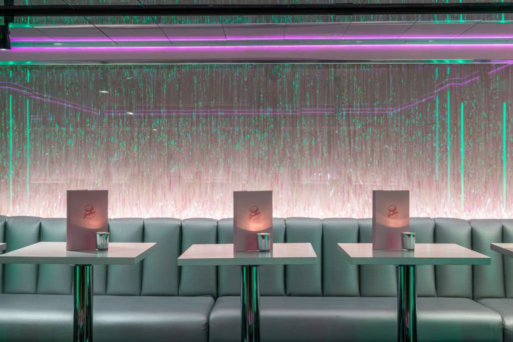 Modern lounge with green banquette seating, individual tables, and neon pink-green wall lighting installation.