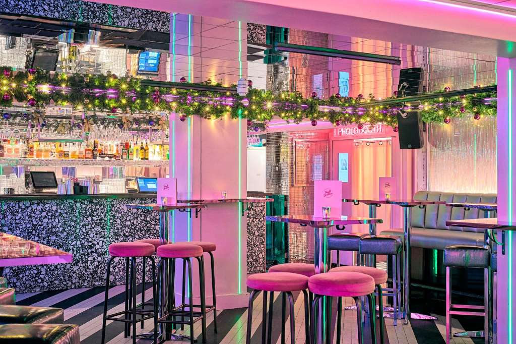 Modern bar and lounge space with pink and neon lighting, bar seating, garland decoration, and contemporary furnishings.