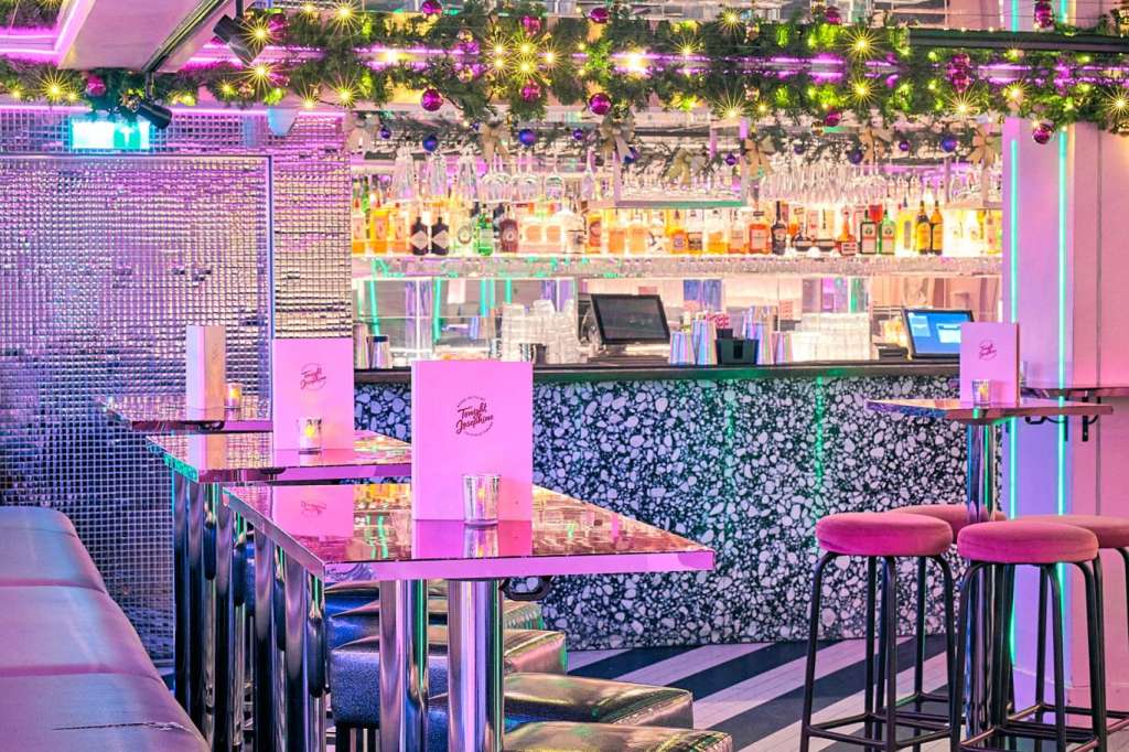 Modern bar and lounge with magenta lighting, high-top tables, bar seating, and decorative floral garland overhead.