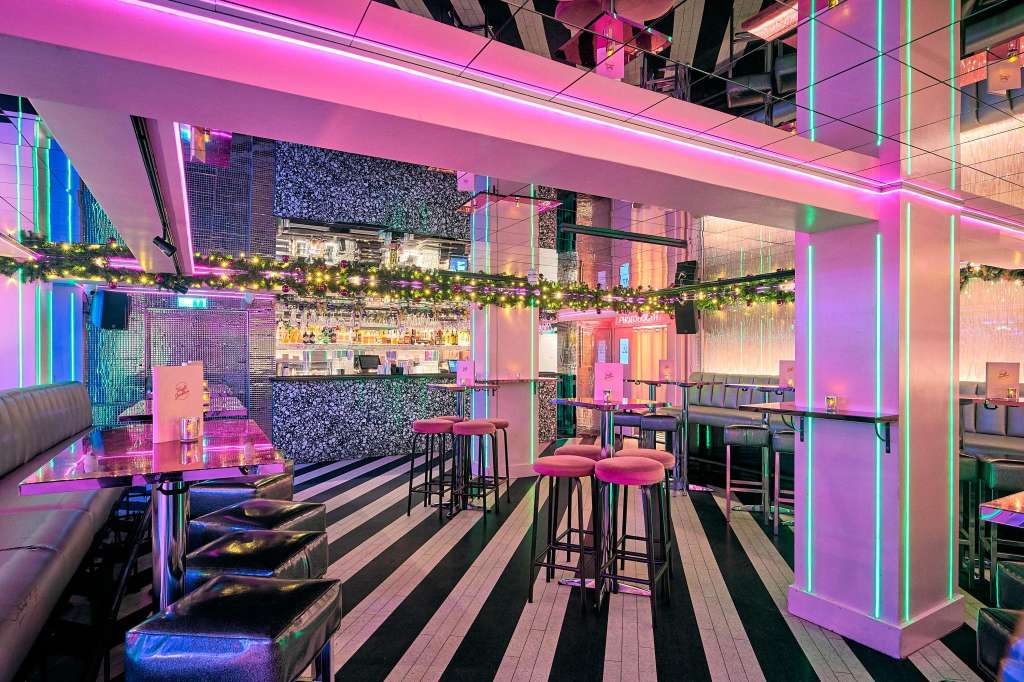 Modern venue interior with neon pink and green lighting, bar counter, high seating, and festive garland decoration.