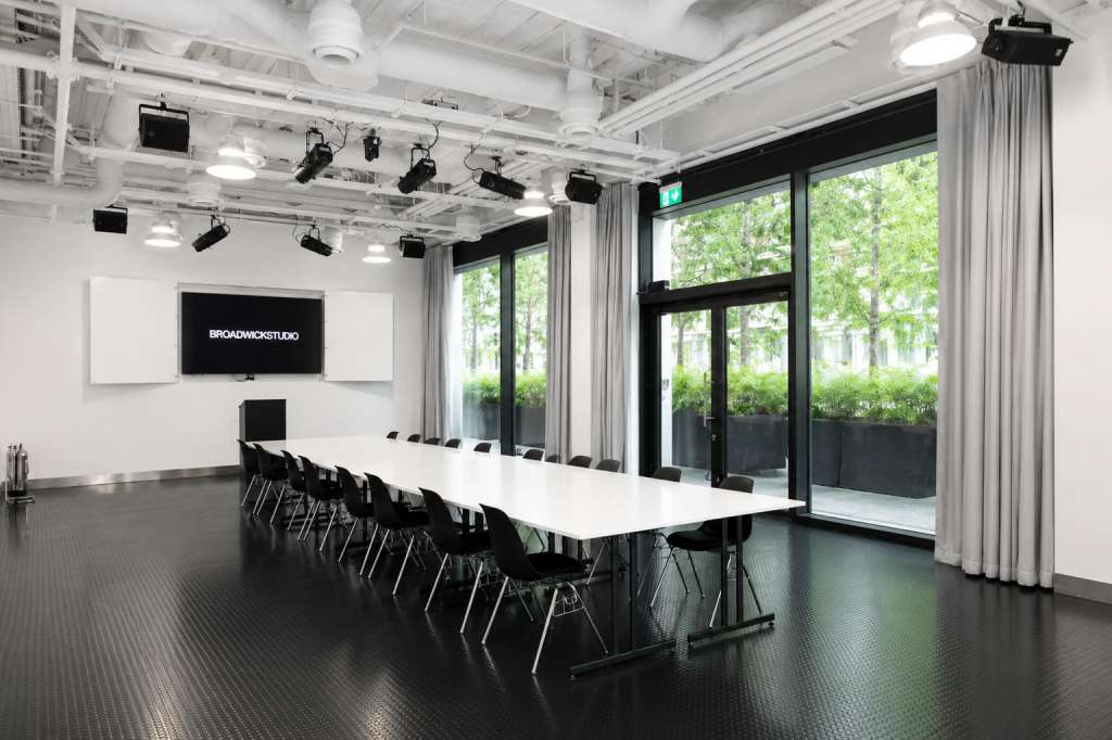 Modern studio space with long white table, black chairs, display screen, professional lighting rig, and floor-to-ceiling windows overlooking greenery.