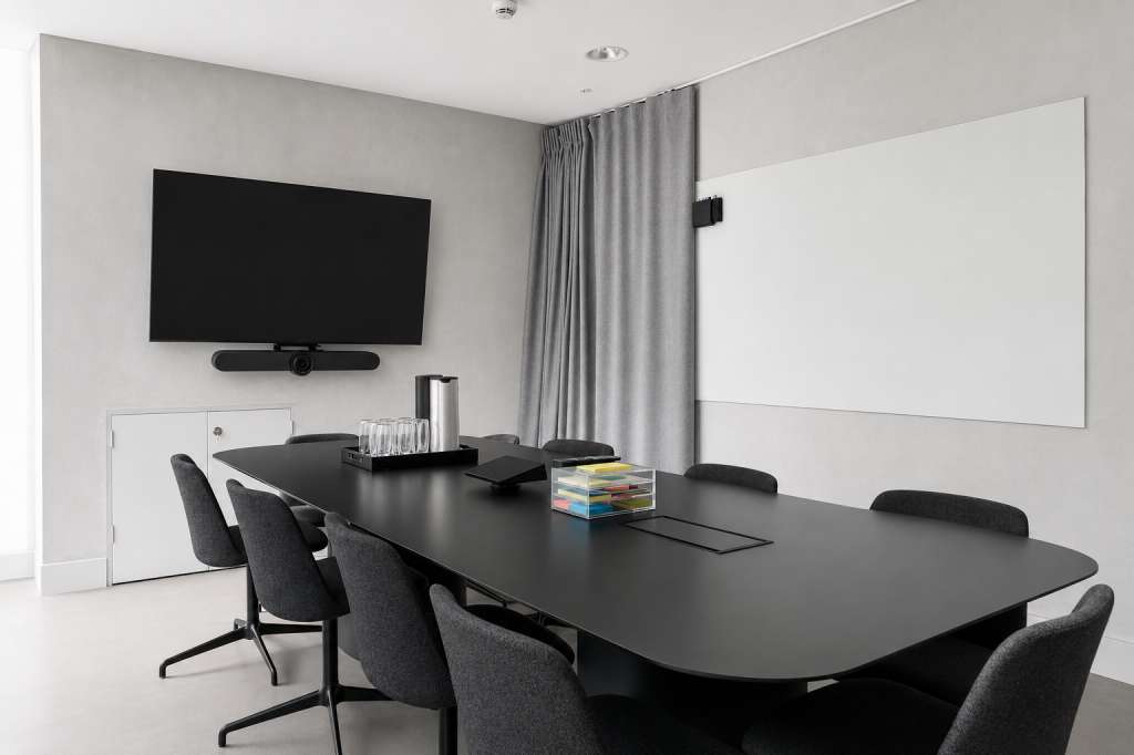 Modern meeting room with long black table, gray chairs, wall-mounted TV, whiteboard, and gray curtains.