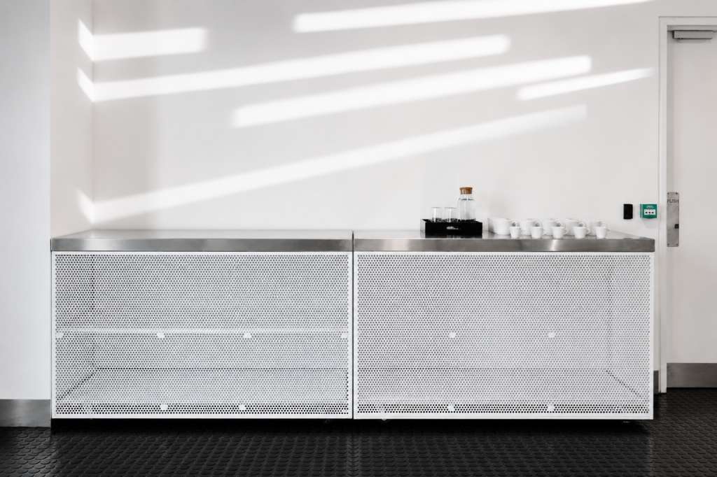 Modern minimalist space with white walls, linear ceiling lights, and a stainless steel counter with perforated white panels.