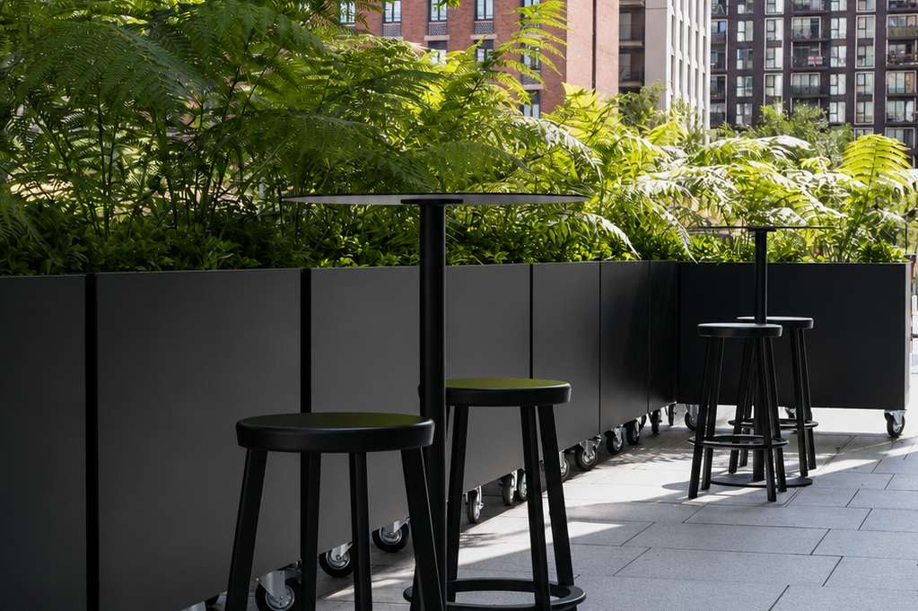 Outdoor terrace with black bar stools, green screening, and lush foliage overlooking urban buildings.