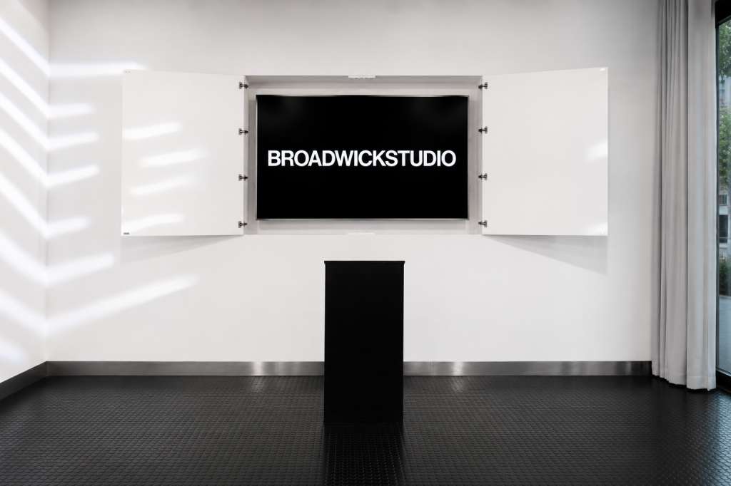 Main space with white walls, black floor, and large display screen showing Broadwick Studio branding.