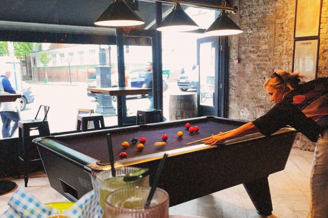 Games room with pool table, pendant lighting, exposed brick walls, and street-facing windows. Person playing pool.