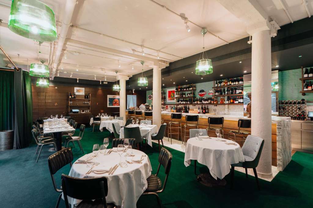 Restaurant dining room with round tables set with white linens, black chairs, green carpet, and bar area with bottle display.